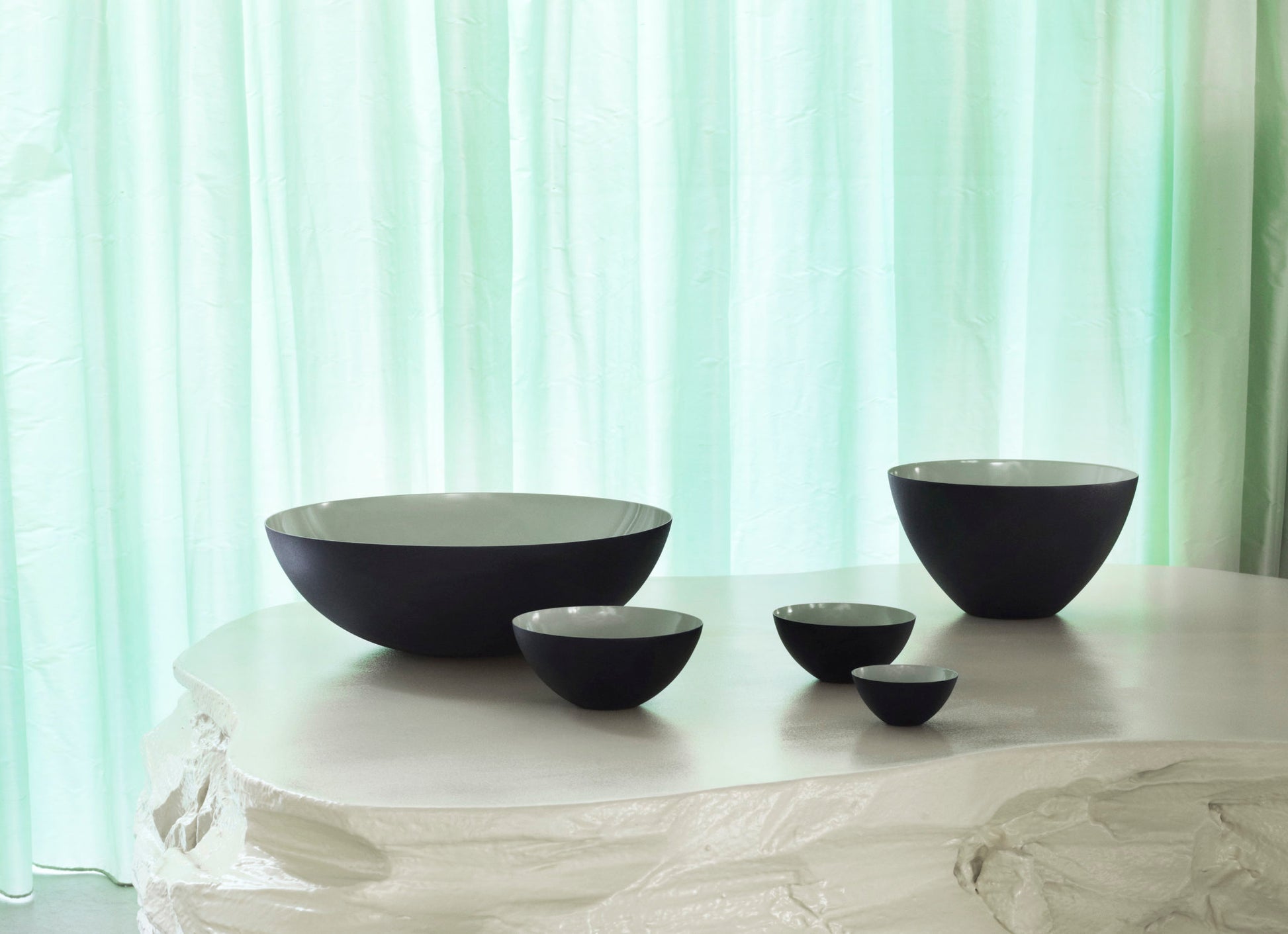 Five Normann Copenhagen Krenit Bowls, 8.4cm in black and white, are arranged on a textured white surface with a light green sheer curtain in the background, reflecting minimalist Danish design.