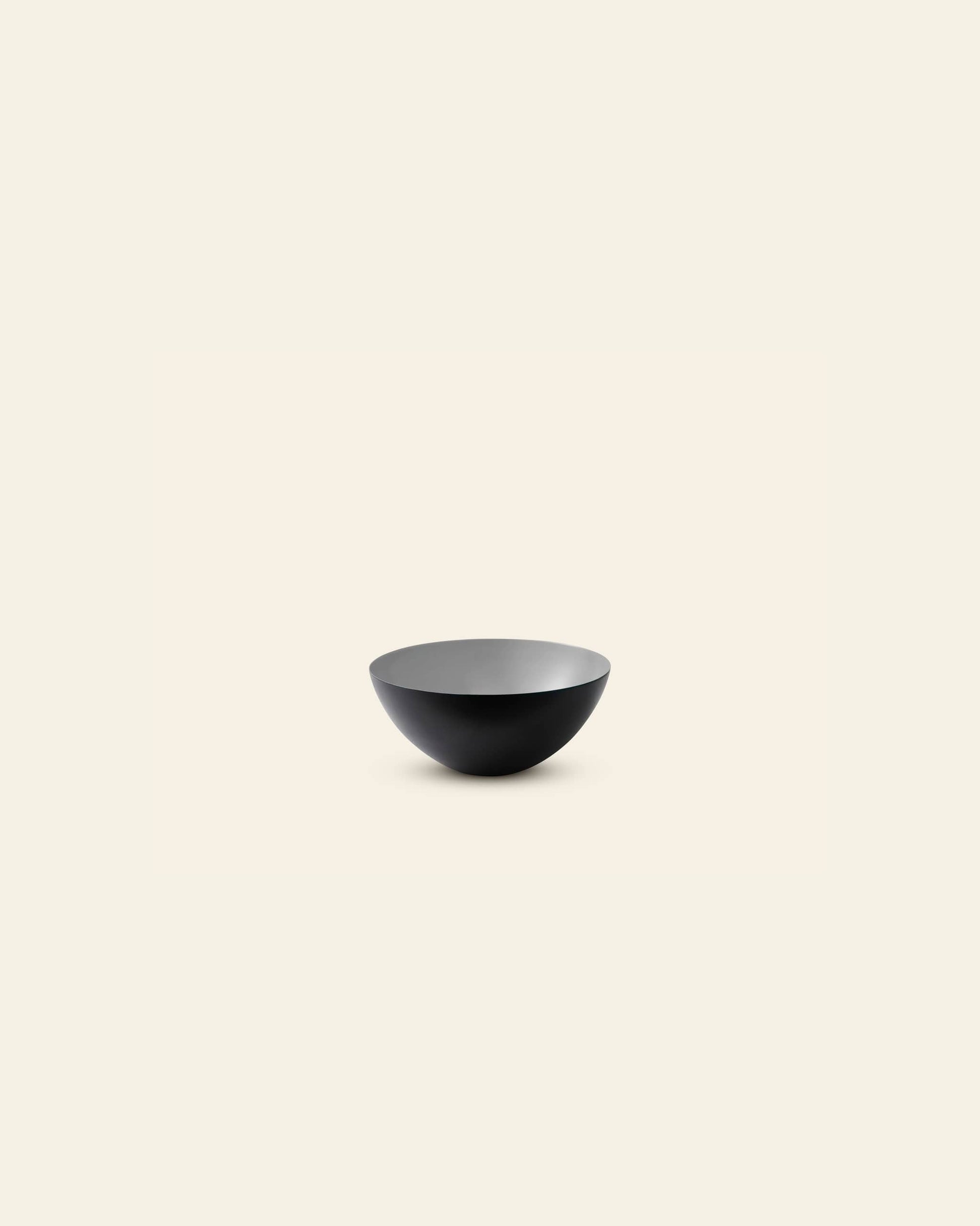 A minimalist image features a single Normann Copenhagen Krenit Bowl, 8.4cm, with a matte black finish, centrally placed on a light cream background. The bowl is empty and viewed from a slight angle, highlighting its classic Danish design.