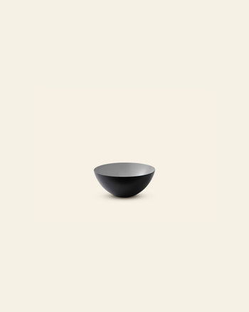 A minimalist image features a single Normann Copenhagen Krenit Bowl, 8.4cm, with a matte black finish, centrally placed on a light cream background. The bowl is empty and viewed from a slight angle, highlighting its classic Danish design.
