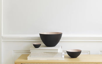 A black matte Normann Copenhagen Krenit Bowl, 8.4cm, sits on a light wooden table alongside white books and decor, exemplifying sleek Danish design against a plain white wall with subtle molding.