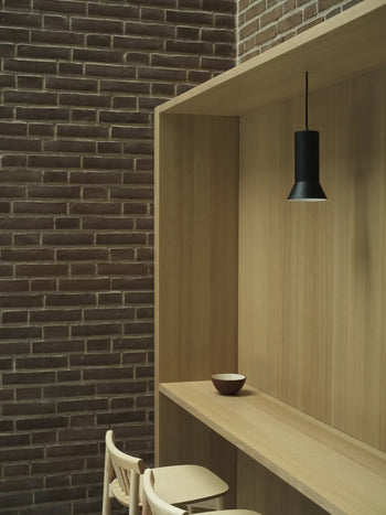 Minimalist wooden nook featuring two light chairs, a narrow counter, and a Normann Copenhagen Line Bowl (15cm) by Normann Copenhagen. A single black pendant light and dark brown brick wall add artistry to the serene space.