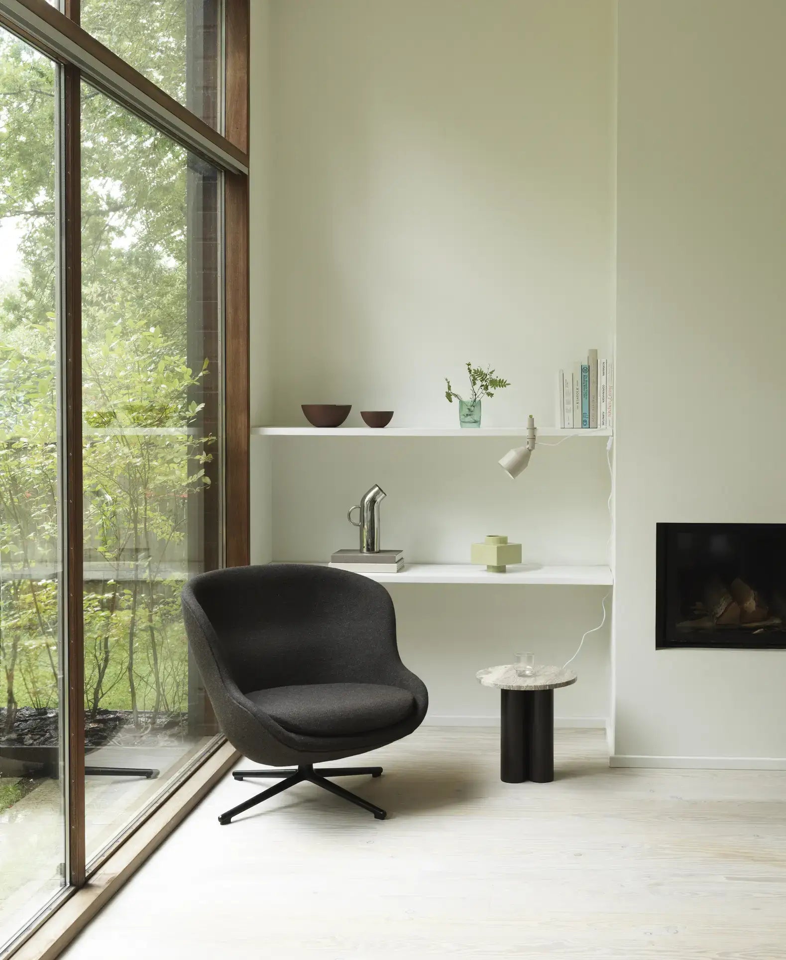 A modern, minimal room with a large window, black swivel chair, white shelves with books and decor, a built-in fireplace, and a Normann Copenhagen Line Bowl (15cm) on a small round side table. Greenery is visible outside the window.