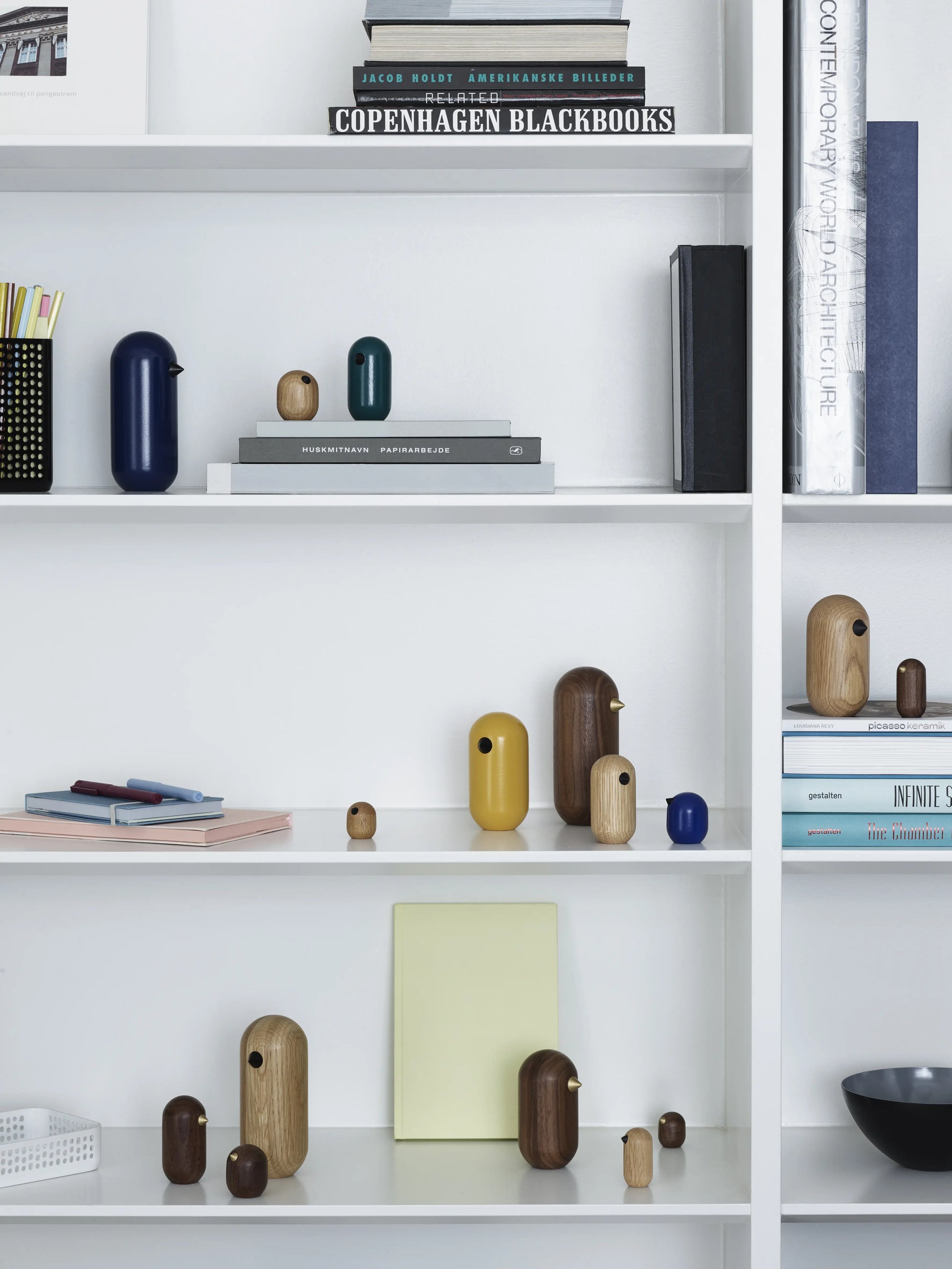 White shelves display neatly arranged books, stacked magazines, and Normann Copenhagens Little Bird (13.5cm) wooden ornaments in blue, yellow, natural wood, and dark brown—adding a decorative touch to this minimalist design.