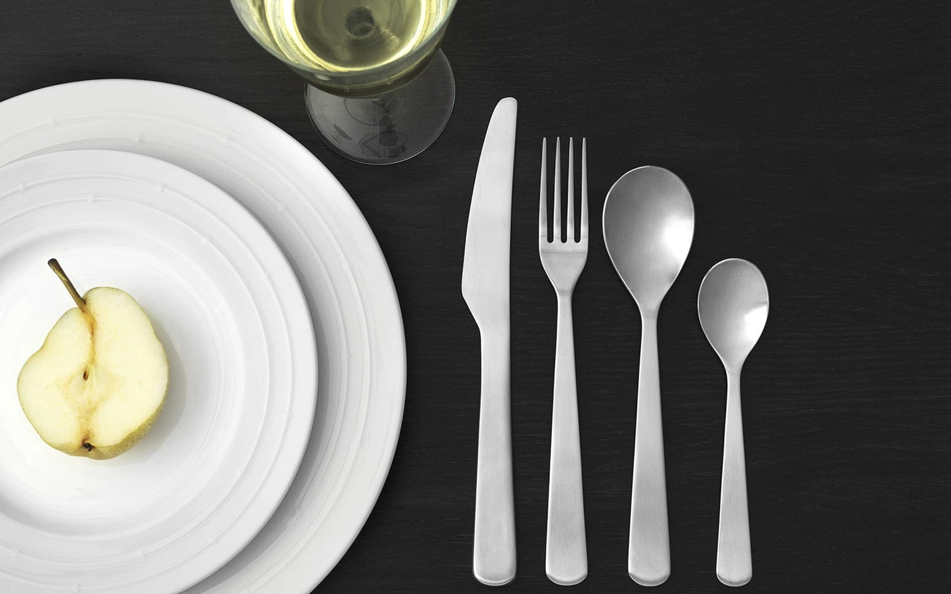 A table setting with stacked white plates holding half a pear, a wine glass, and the Normann Copenhagen Cutlery Gift Box—16 polished modern pieces (knife, fork, tablespoon, teaspoon)—by Normann Copenhagen on a dark surface.