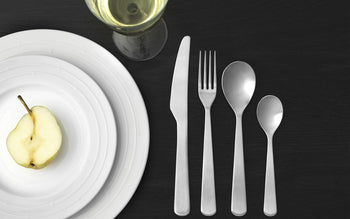 A table setting with stacked white plates holding half a pear, a wine glass, and the Normann Copenhagen Cutlery Gift Box—16 polished modern pieces (knife, fork, tablespoon, teaspoon)—by Normann Copenhagen on a dark surface.