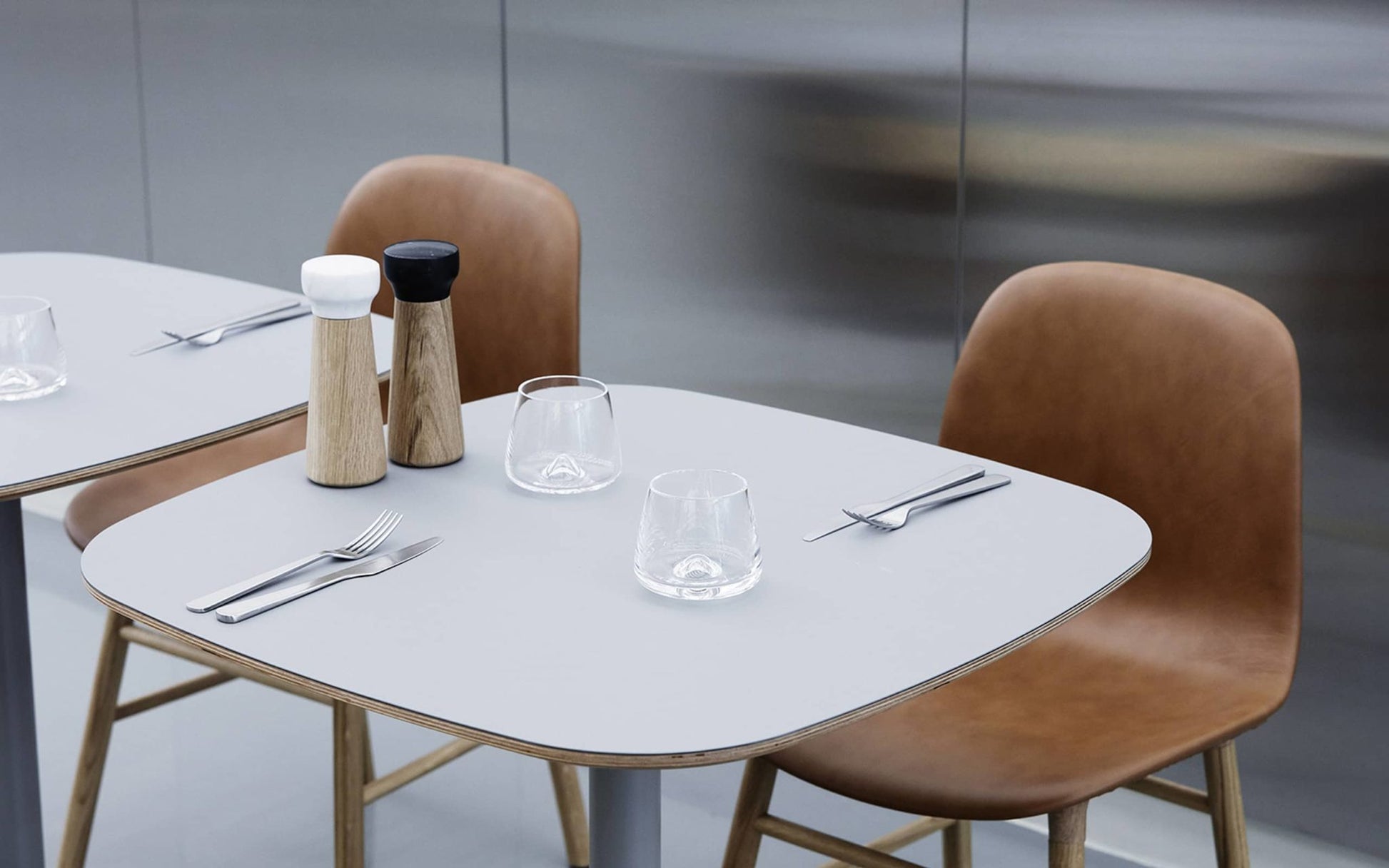 Two modern dining tables with brown chairs and a polished finish, each set with Normann Copenhagen Cutlery Gift Box (16 Pack), two glasses, and salt & pepper shakers, against a metallic wall background.