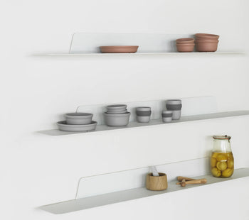 Three minimalist white wall shelves display stacked bowls, cups, and containers in neutral tones, including the Normann Copenhagen Obi Mug by Normann Copenhagen. A glass jar with yellow contents and a wooden honey dipper sit on the bottom shelf.