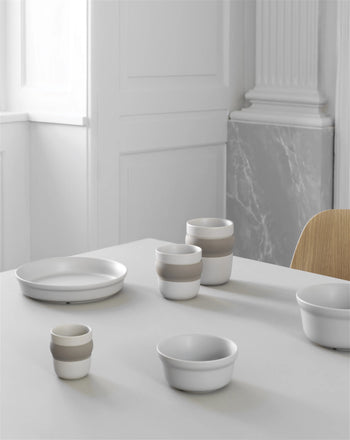 A minimalist dining table set with white and beige ceramic bowls, cups, a plate, and the Normann Copenhagen Obi Mug. In the background are white paneled walls, a marble accent, and part of a light wooden chair.