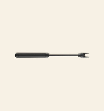 The Normann Copenhagen Pebble Cheese Fork, a black two-pronged fork with a long handle, is centered on a plain light beige background.