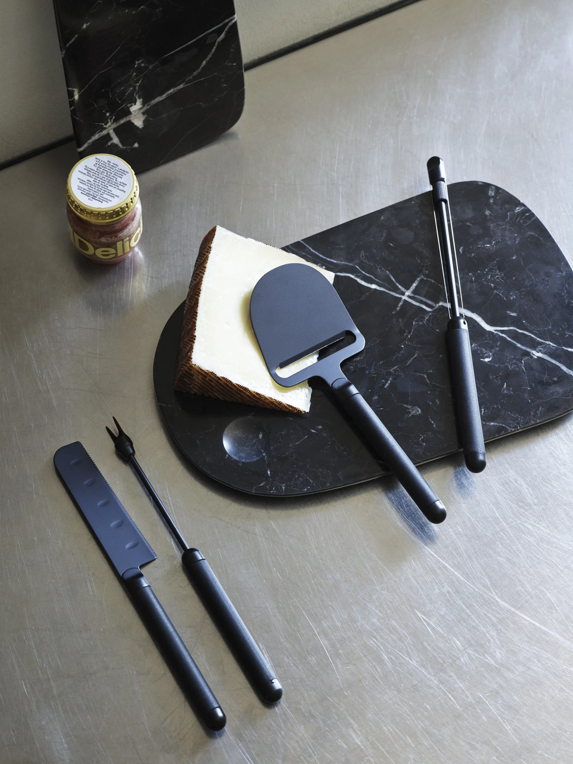 A wedge of cheese sits on a black marble board with a slicer. Nearby are a Normann Copenhagen Pebble Cheese Fork, a cheese knife, metal tongs, and a jar of jam on a stainless steel surface.