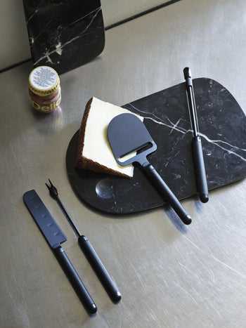 A wedge of cheese sits on a black marble board with a slicer. Nearby are a Normann Copenhagen Pebble Cheese Fork, a cheese knife, metal tongs, and a jar of jam on a stainless steel surface.