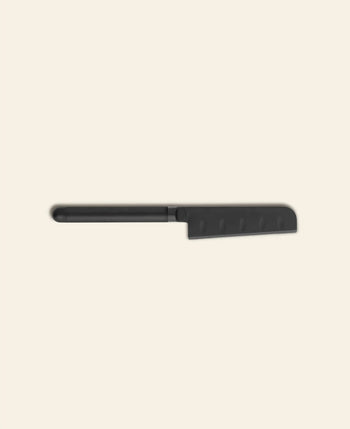 The Normann Copenhagen Pebble Cheese Knife by Normann Copenhagen features a black, serrated blade with a straight handle and rounded tip, inspired by Scandinavian design, shown on a light beige background.