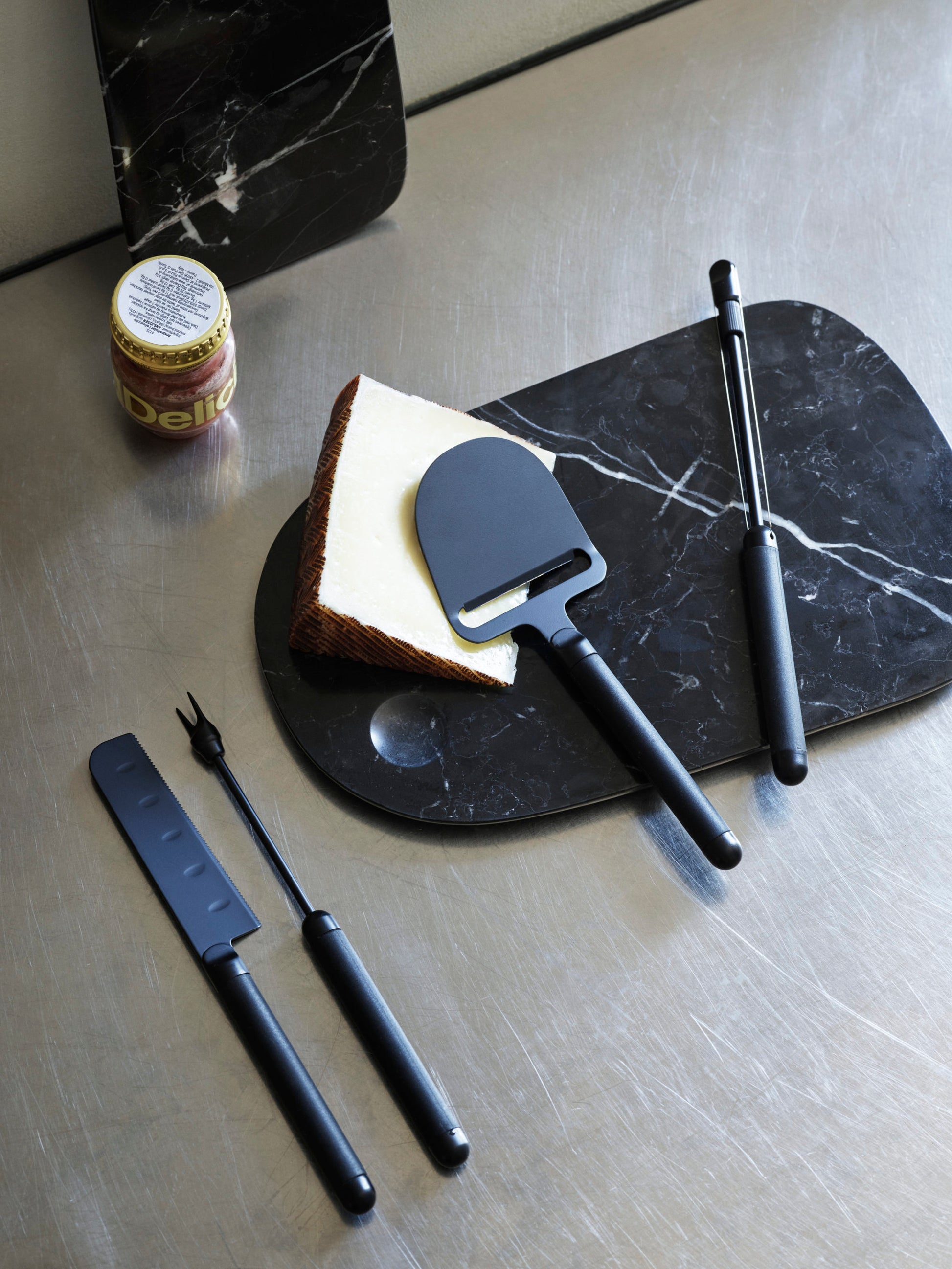 The Normann Copenhagen Pebble Cheese Knife sits with cheese and accessories on a black marble board, exemplifying Scandinavian design—perfect for modern hosts—next to a jar of Delice spread on a stainless steel countertop.