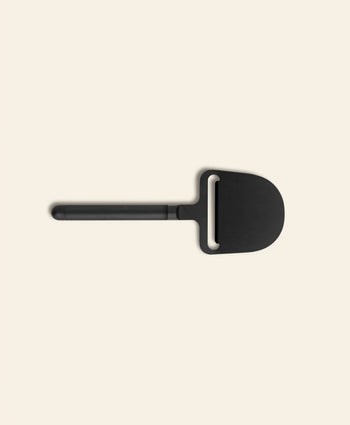 The Normann Copenhagen Pebble Cheese Plane by Normann Copenhagen showcases a sleek black, wide blade and straight handle, ideal for smooth cheese serving, displayed on a plain light beige background.