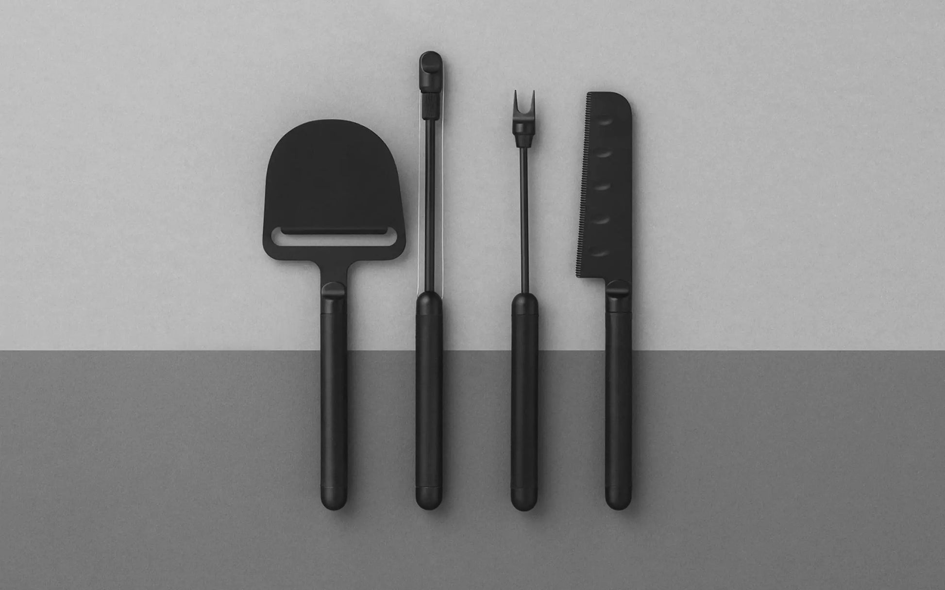 Four black cheese utensils—including a slicer, spreader, fork, and the Normann Copenhagen Pebble Cheese Plane—are neatly lined up on a two-tone gray background, ideal for stylish cheese serving.