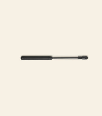 A black and silver gas strut, echoing the sleek look of the Normann Copenhagen Pebble Cheese Slicer, lies horizontally on a light beige background with its thicker black end to the left.