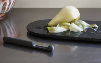 A partially peeled pear sits on a black cutting board with strips of peel beside it. In the foreground, a Normann Copenhagen Pebble Cheese Slicer rests on a stainless steel countertop.