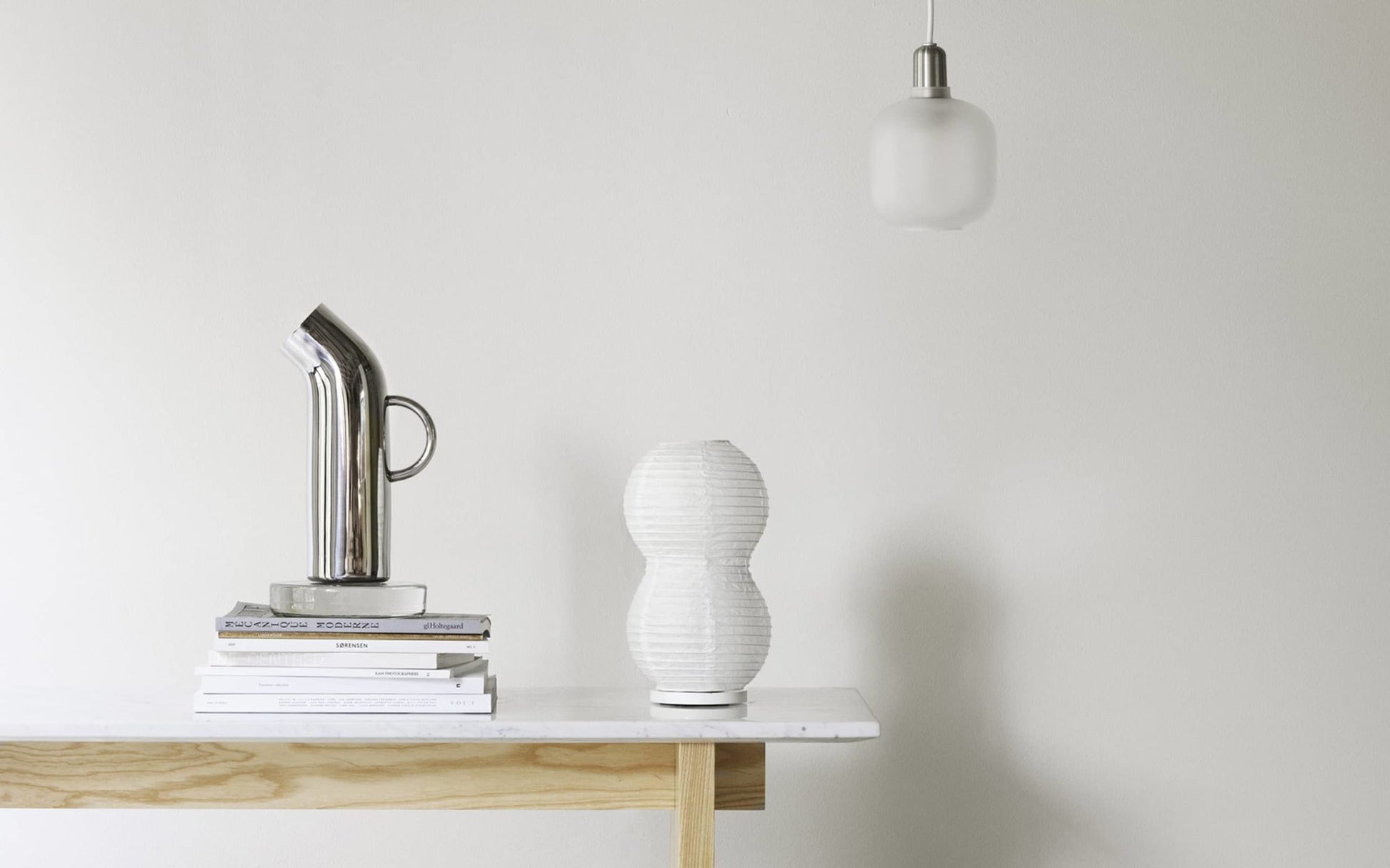 A minimalist scene with a light wooden table displaying a stack of books, the Normann Copenhagen Pipe Pitcher in retro silver, and a white textured vase. A frosted glass pendant light hangs above against a plain white wall.