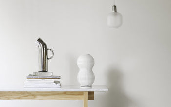 A minimalist scene with a light wooden table displaying a stack of books, the Normann Copenhagen Pipe Pitcher in retro silver, and a white textured vase. A frosted glass pendant light hangs above against a plain white wall.