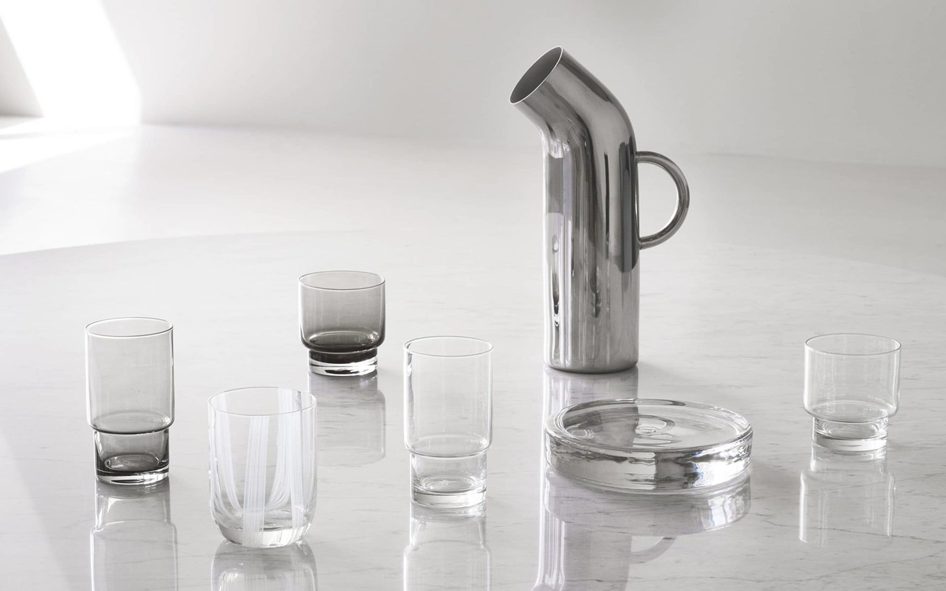 A sleek Normann Copenhagen Pipe Pitcher with five clear and smoky glasses and a round glass tray sits on a glossy marble table, adding subtle retro style to your kitchen. The minimalistic background enhances its modern appeal.