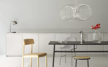 A modern dining area with a black table, three chairs, a Normann Copenhagen Pipe Pitcher, a glass of water, red flowers in a vase, a desk lamp, and a sculptural pendant light in a neutral-toned kitchen.