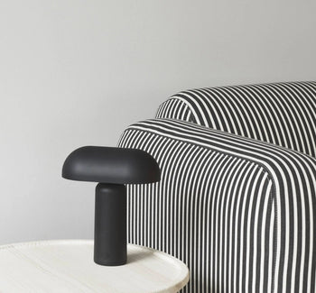 A Normann Copenhagen Porta Table Lamp with a modern design sits on a light-colored round table beside a sofa with black and white stripes, set against a plain light gray wall.