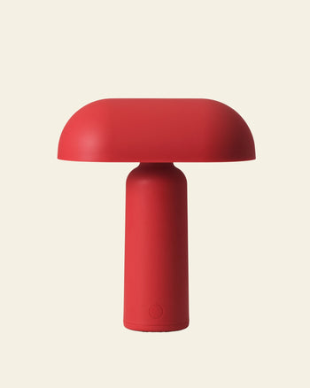 The Normann Copenhagen Porta Table Lamp by Normann Copenhagen is a modern, minimalist red lamp featuring a cylindrical base and rounded dome top, shown against a plain light beige background.