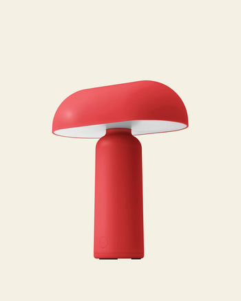 The Normann Copenhagen Porta Table Lamp is a modern, red portable lamp with a cylindrical base and wide, mushroom-like shade—sleekly designed by Normann Copenhagen and set against a light beige background.