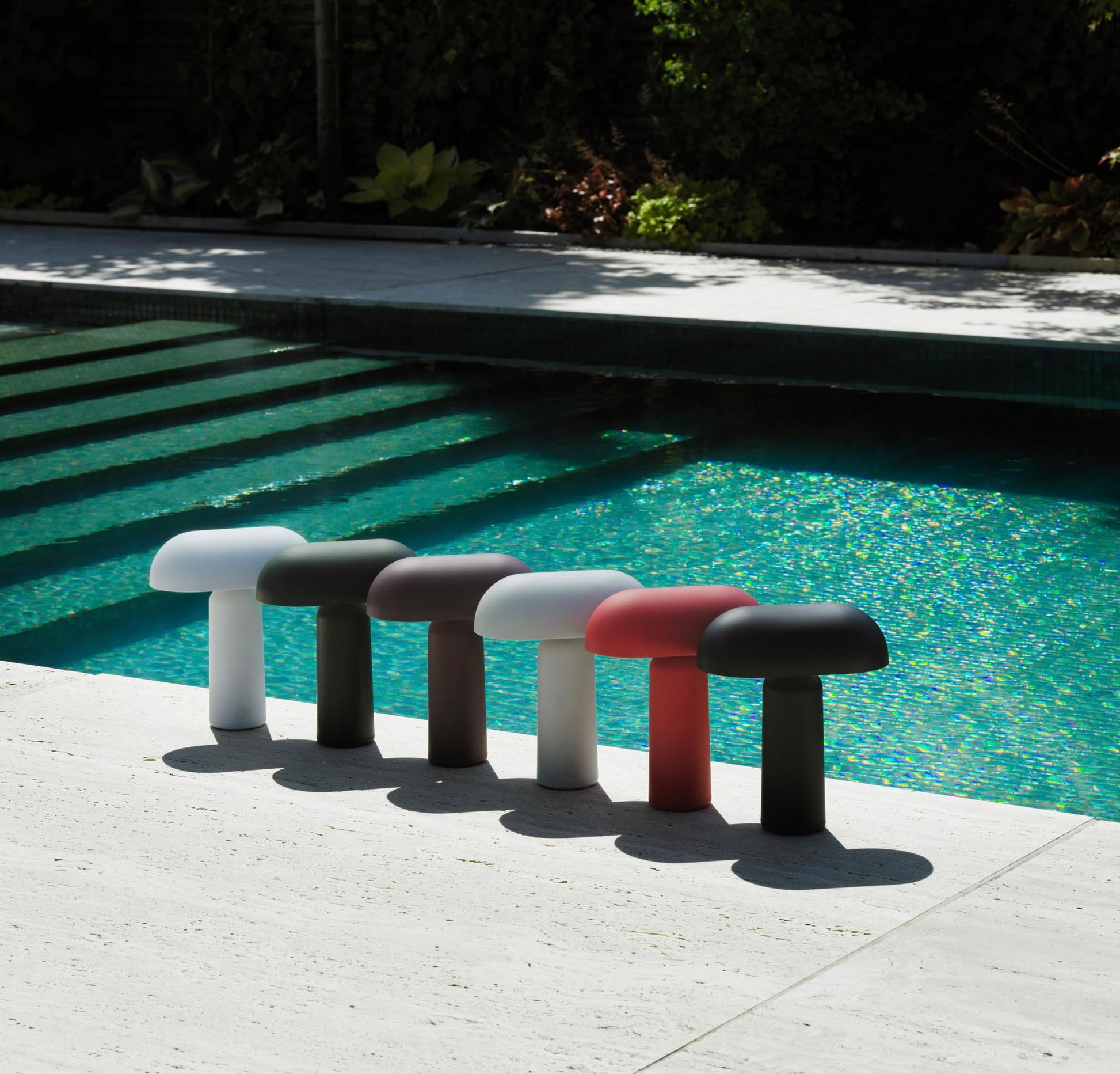 Six Normann Copenhagen Porta Table Lamps, with modern design, are lined up by a sunlit pool with clear turquoise water. Light and shadow play across the bright stone poolside, highlighting their stylish presence.