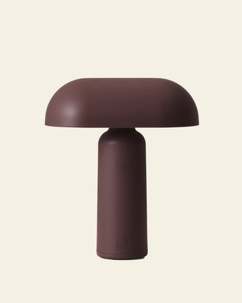 The Normann Copenhagen Porta Table Lamp features a minimalist design with a matte brown finish, cylindrical base, and wide dome-shaped shade, inspired by Modern Design. Shown against an off-white background.