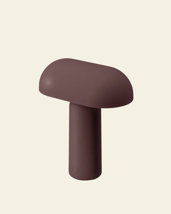 The Normann Copenhagen Porta Table Lamp by Normann Copenhagen is a brown, modern, mushroom-shaped portable lamp with a rounded top and cylindrical base, shown against a plain light beige background.
