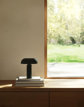 A sleek black Normann Copenhagen Porta Table Lamp rests on a stack of books by a large window, sunlight brightening the minimal space and offering a view of grass and trees outside.