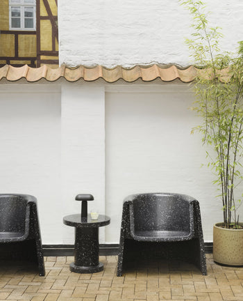 Two black speckled armchairs and a Normann Copenhagen Porta Table Lamp on a matching side table sit by a white wall with tiled roof. To the right is a tall potted bamboo, while brick paving covers the modern floor.