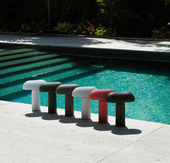 Six colorful Normann Copenhagen Porta Table Lamps from Normann Copenhagen are arranged on a light stone poolside, their modern design and portable style casting distinct shadows against the blue pool water and lush greenery.