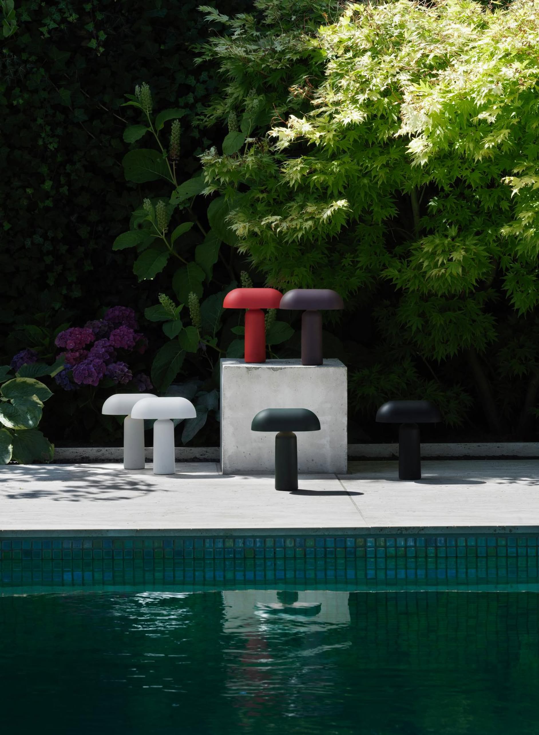 Five Normann Copenhagen Porta Table Lamps in white, red, brown, black, and dark green sit poolside on concrete, their modern design standing out amid lush greenery and purple flowers.