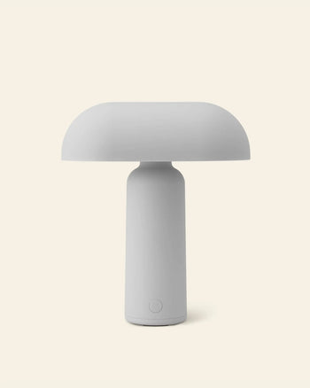 The Normann Copenhagen Porta Table Lamp features a minimalist, modern design with a mushroom-shaped shade and cylindrical base, showcased against a plain, light beige background.