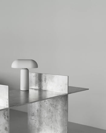 The Normann Copenhagen Porta Table Lamp, featuring a minimalist white dome-shaped shade, sits on a metallic industrial table against a plain light gray background, highlighting the modern style of Normann Copenhagen.