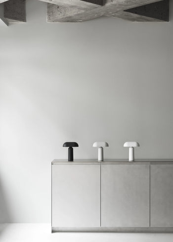 A minimalist room features a concrete ceiling, light gray walls and cabinets, and three Normann Copenhagen Porta Table Lamps in black, light gray, and white arranged on top of the cabinet.