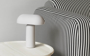 The Normann Copenhagen Porta Table Lamp in white sits on a round, light-colored table beside a black-and-white striped chair, showcasing minimalist, contemporary design by Normann Copenhagen.