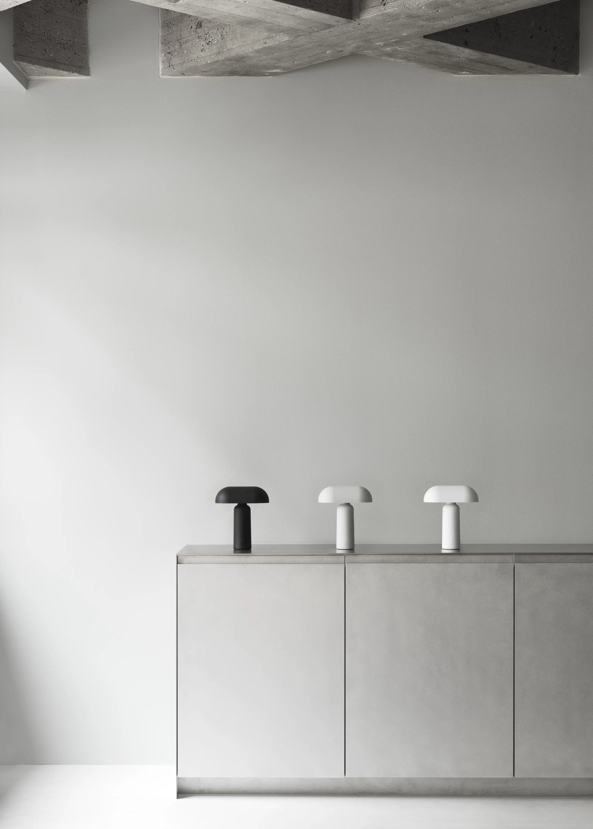 A minimalist room showcases a light gray cabinet topped with three Normann Copenhagen Porta Table Lamps in black, gray, and white. Above, a textured concrete ceiling contrasts with plain, light-colored walls.