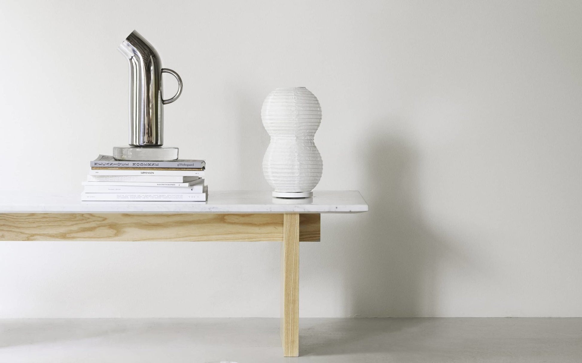 A minimalist table with light wood legs and a white top holds a stack of books, a silver pitcher, and the Normann Copenhagen Puff Table Lamp Twist—a modern fixture—against a plain light-colored wall.