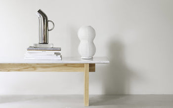 A minimalist table with light wood legs and a white top holds a stack of books, a silver pitcher, and the Normann Copenhagen Puff Table Lamp Twist—a modern fixture—against a plain light-colored wall.