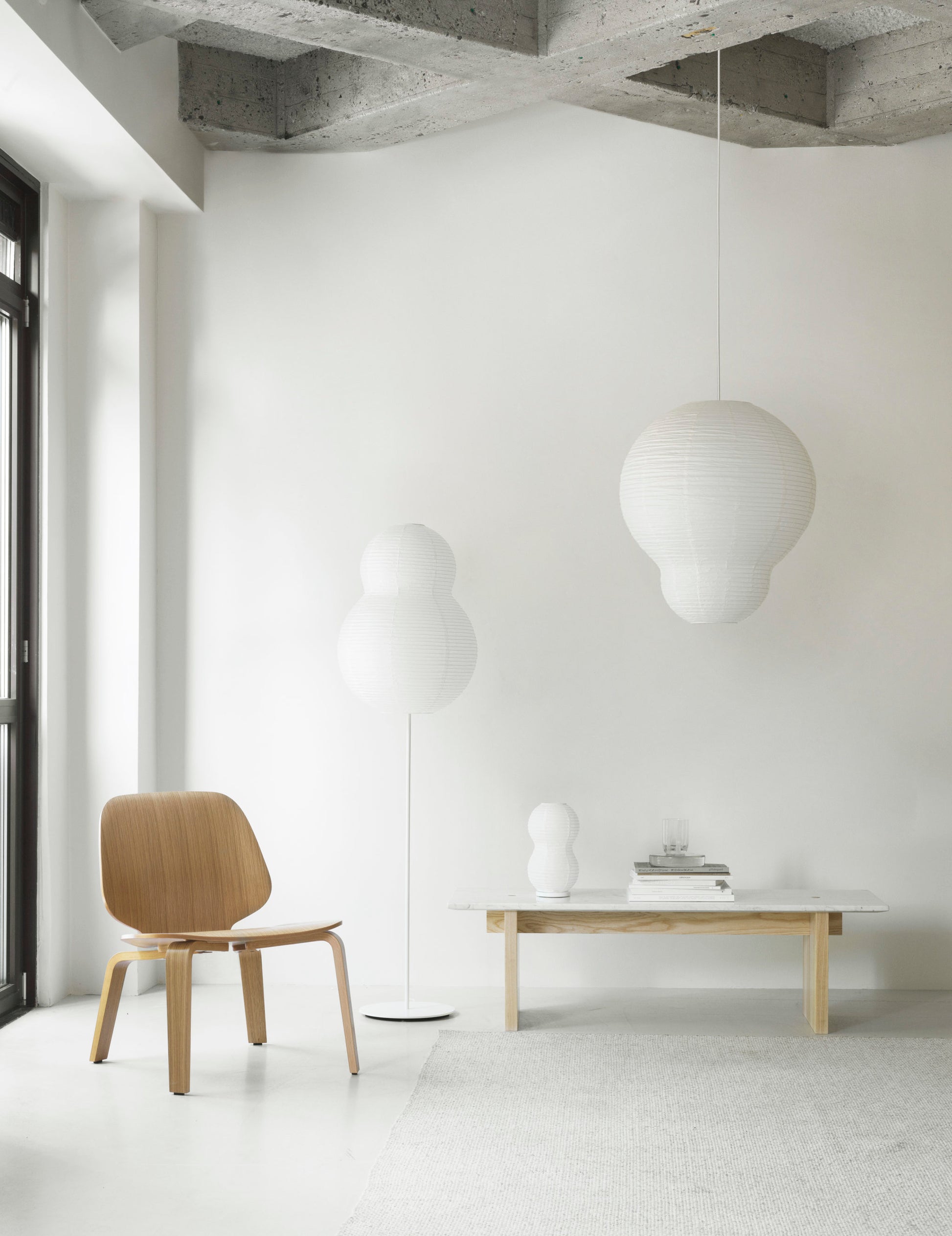 A minimalist room features a wooden chair, low table, and the Normann Copenhagen Puff Table Lamp Twist as a modern light. A hanging paper lantern accents the neutral decor, with natural light streaming in from the left onto white walls and a concrete ceiling.