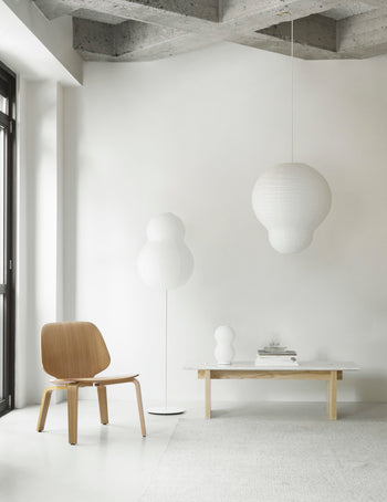 A minimalist room features a wooden chair, low table, and the Normann Copenhagen Puff Table Lamp Twist as a modern light. A hanging paper lantern accents the neutral decor, with natural light streaming in from the left onto white walls and a concrete ceiling.