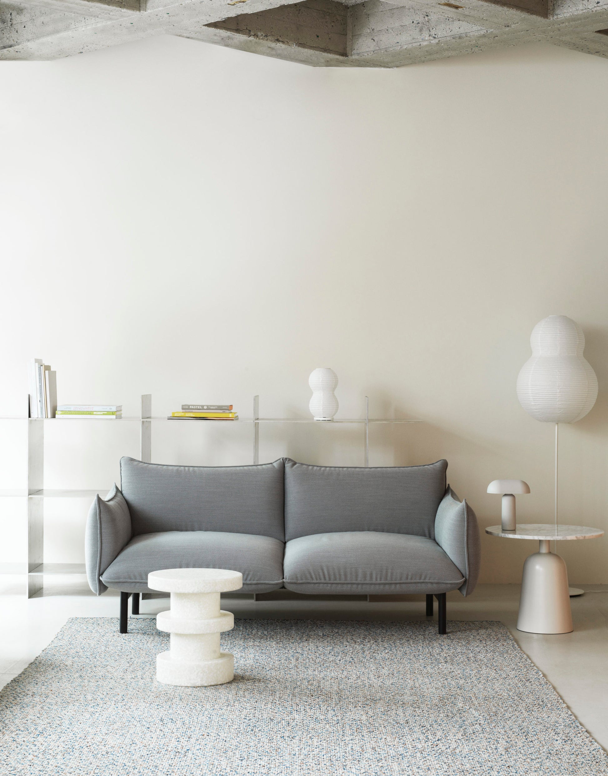 A modern living room with a light gray sofa, small round textured coffee table, and minimalist floor lamp. A side table displays the Normann Copenhagen Puff Table Lamp Twist, bringing unique modern lighting to the neutral decor against a cream wall.