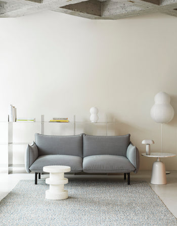 A modern living room with a light gray sofa, small round textured coffee table, and minimalist floor lamp. A side table displays the Normann Copenhagen Puff Table Lamp Twist, bringing unique modern lighting to the neutral decor against a cream wall.