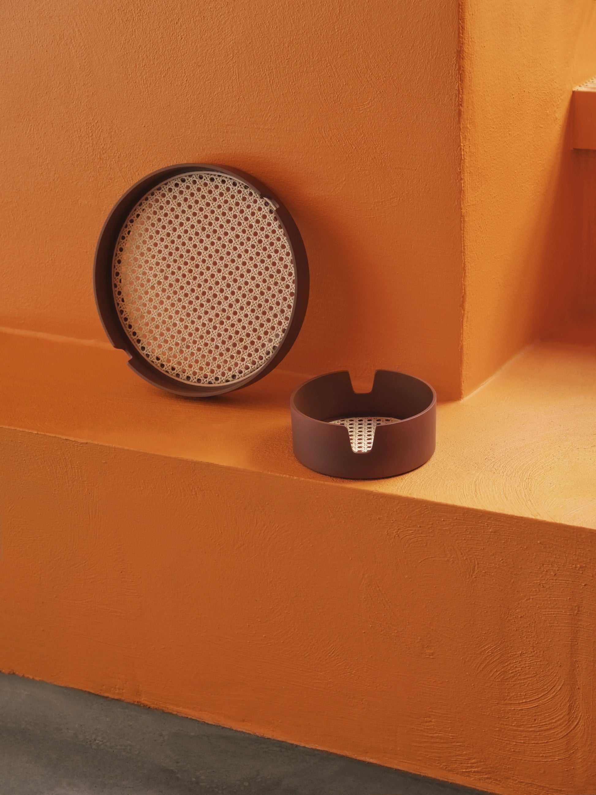 Two modern round brown trays with woven beige inserts, the Normann Copenhagen Salon Tray (40cm), are displayed against a textured orange wall and step—one upright, the other lying flat.
