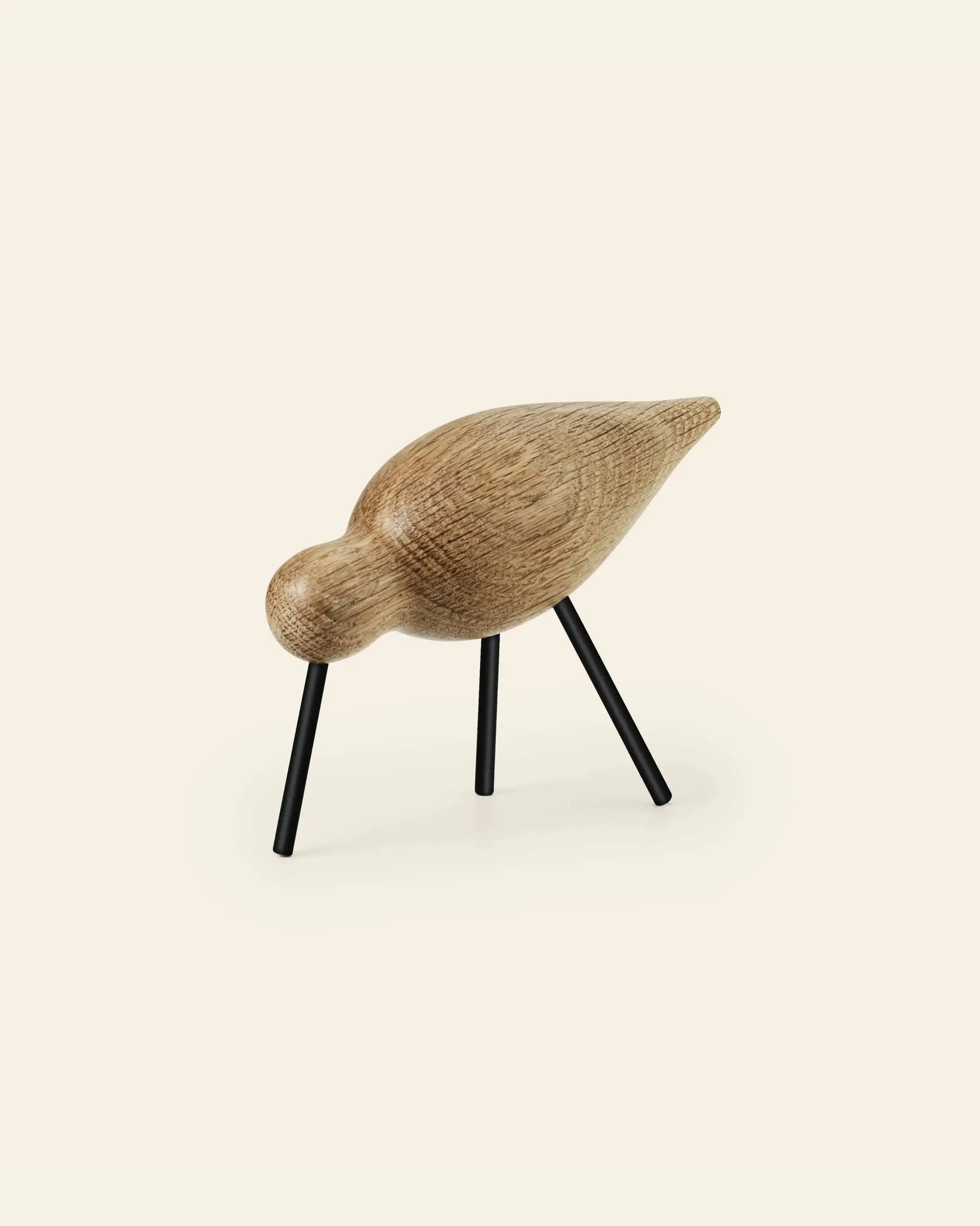 The Normann Copenhagen Shorebird Medium Oak by Normann Copenhagen is a minimalist oak wood bird sculpture with a smooth, rounded body, slim black legs, and is displayed against an off-white background.