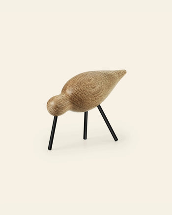 The Normann Copenhagen Shorebird Medium Oak by Normann Copenhagen is a minimalist oak wood bird sculpture with a smooth, rounded body, slim black legs, and is displayed against an off-white background.