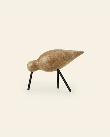 The Normann Copenhagen Shorebird Medium Oak is a minimalist oak wood bird sculpture with a rounded body, long pointed tail, and two black metal legs, inspired by Normann Copenhagen’s iconic design.
